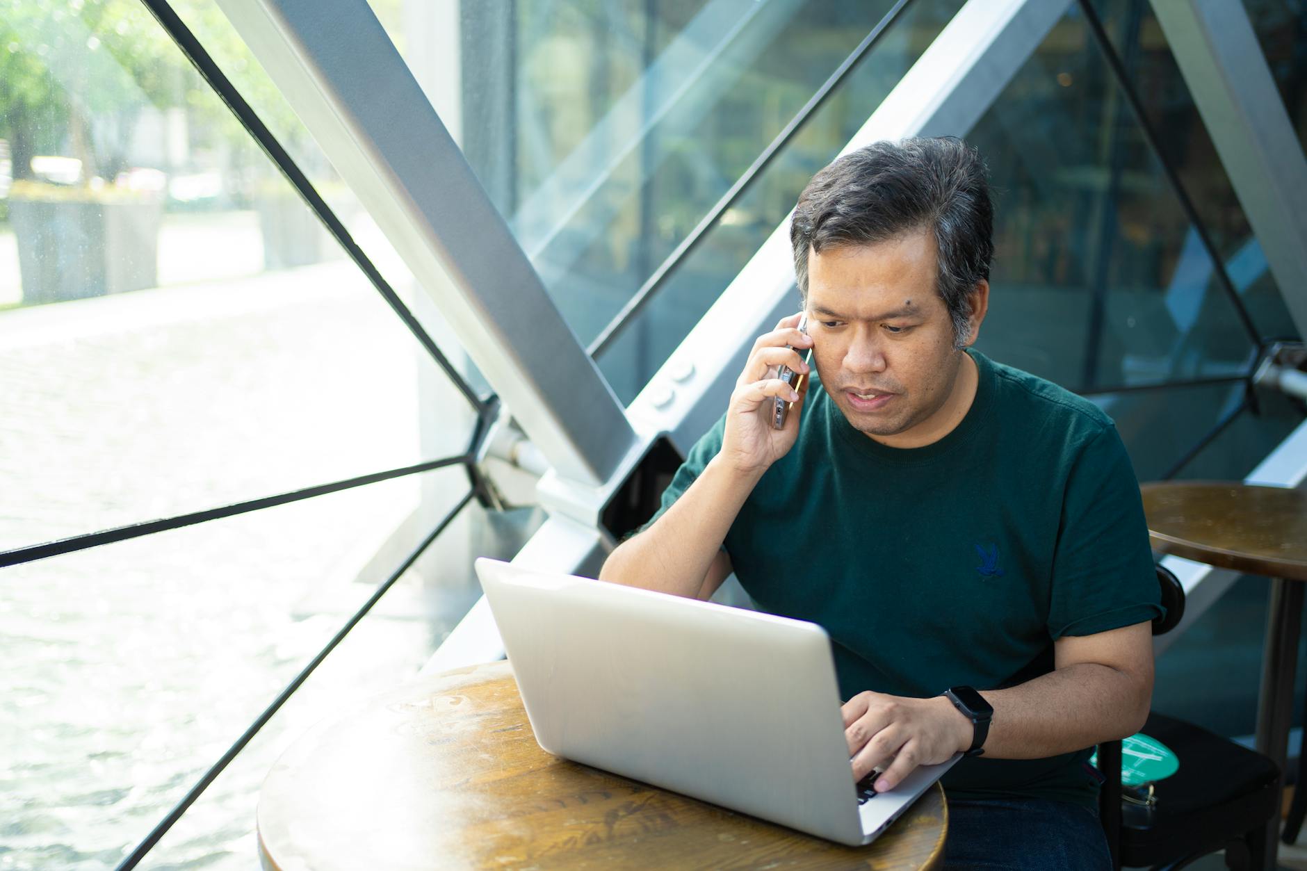A man on a phone call with an open laptop at a table beside a glass wall