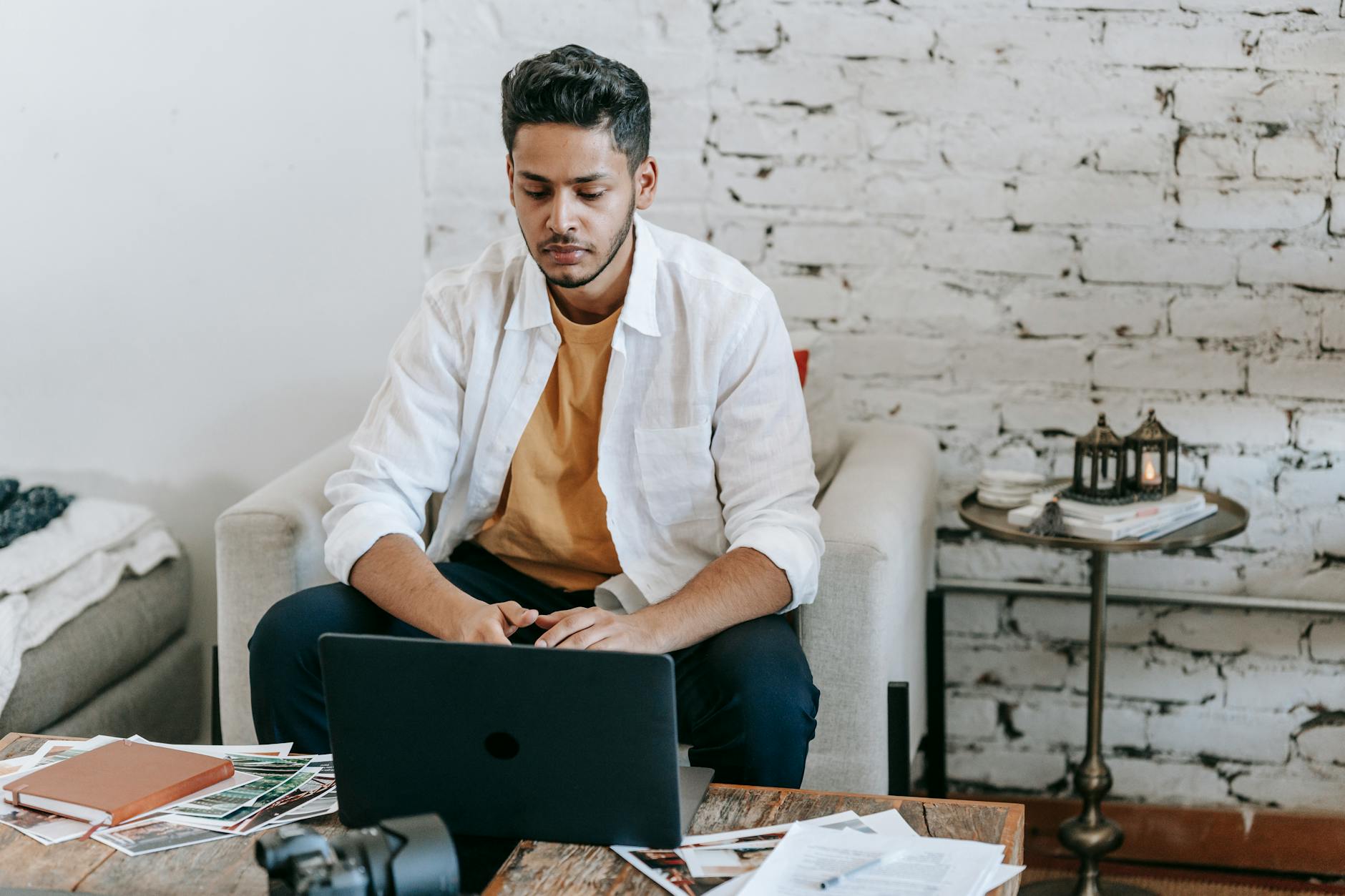 A man at a laptop in an armchair with notebooks and printed photos on a coffee table