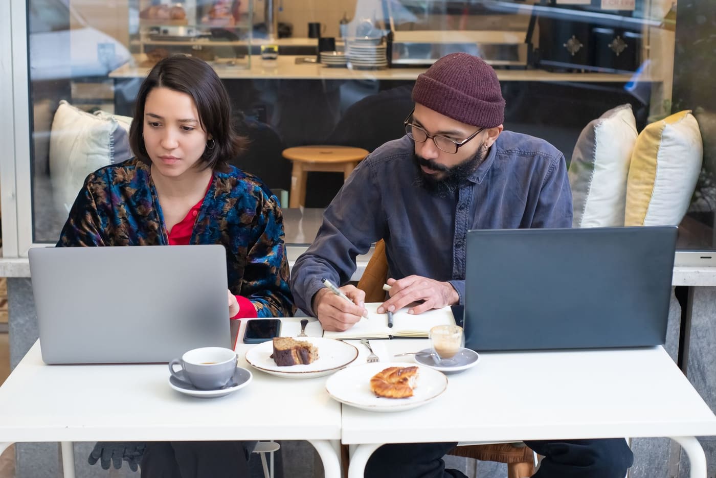 Two people with laptops, coffee and pastries at a cafe table, one taking notes