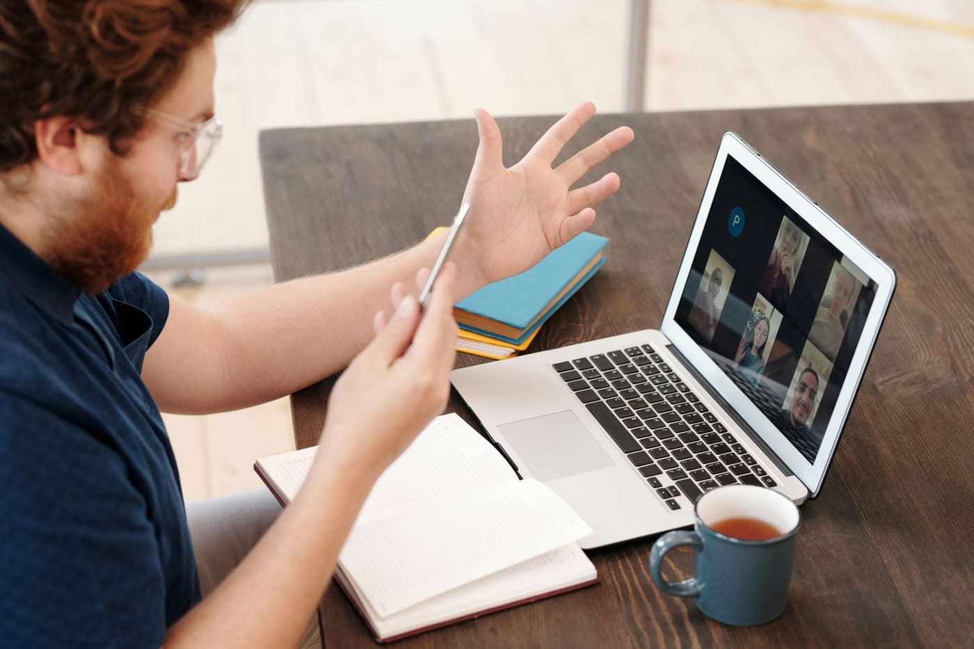 A man gesturing with a notebook in hand during a video call on a laptop