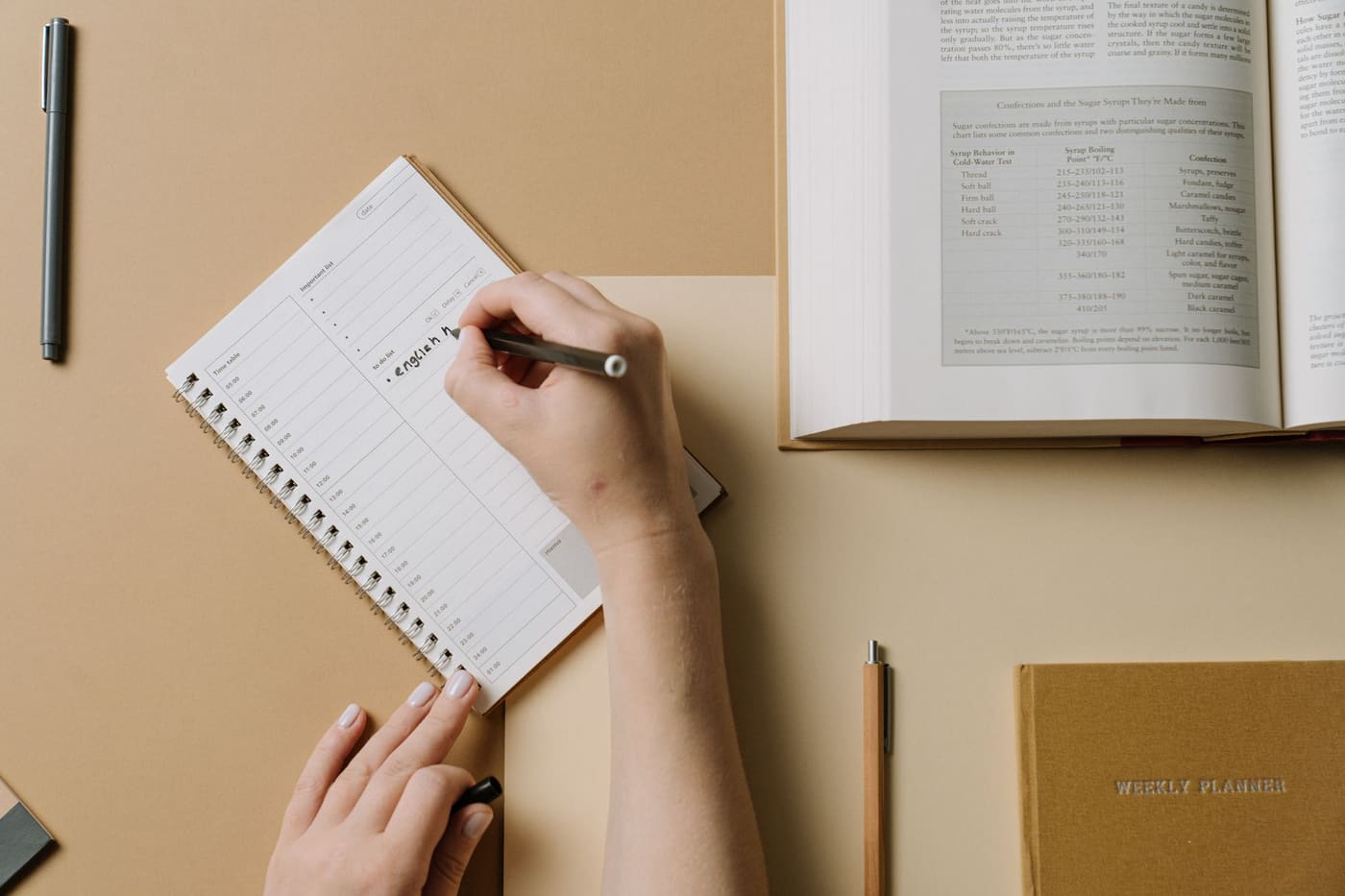 Hands writing in a planner at a desk with an open book nearby