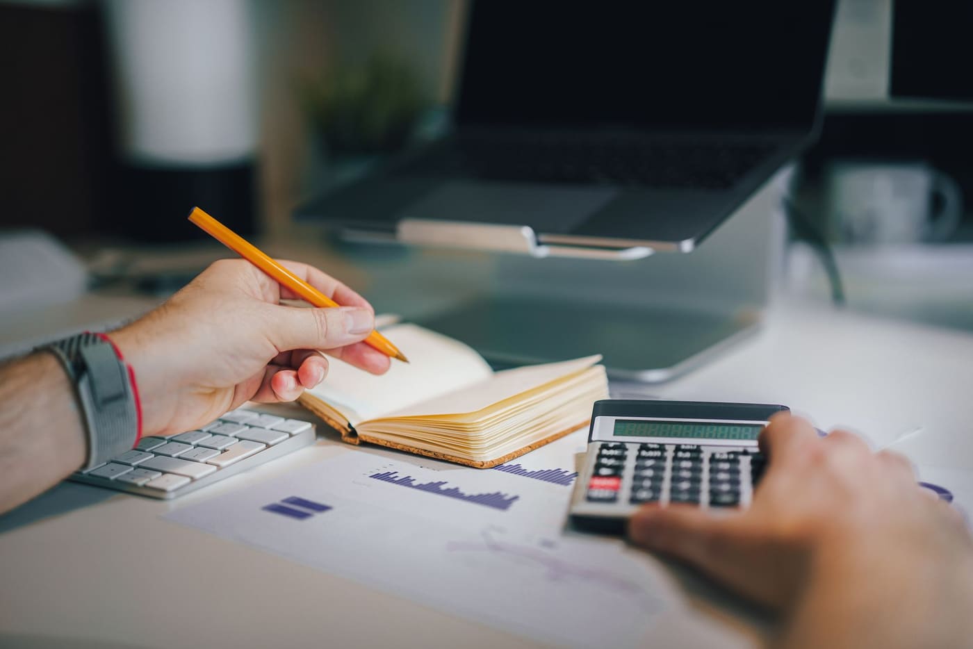 Hands using a calculator and taking notes at a desk with a laptop