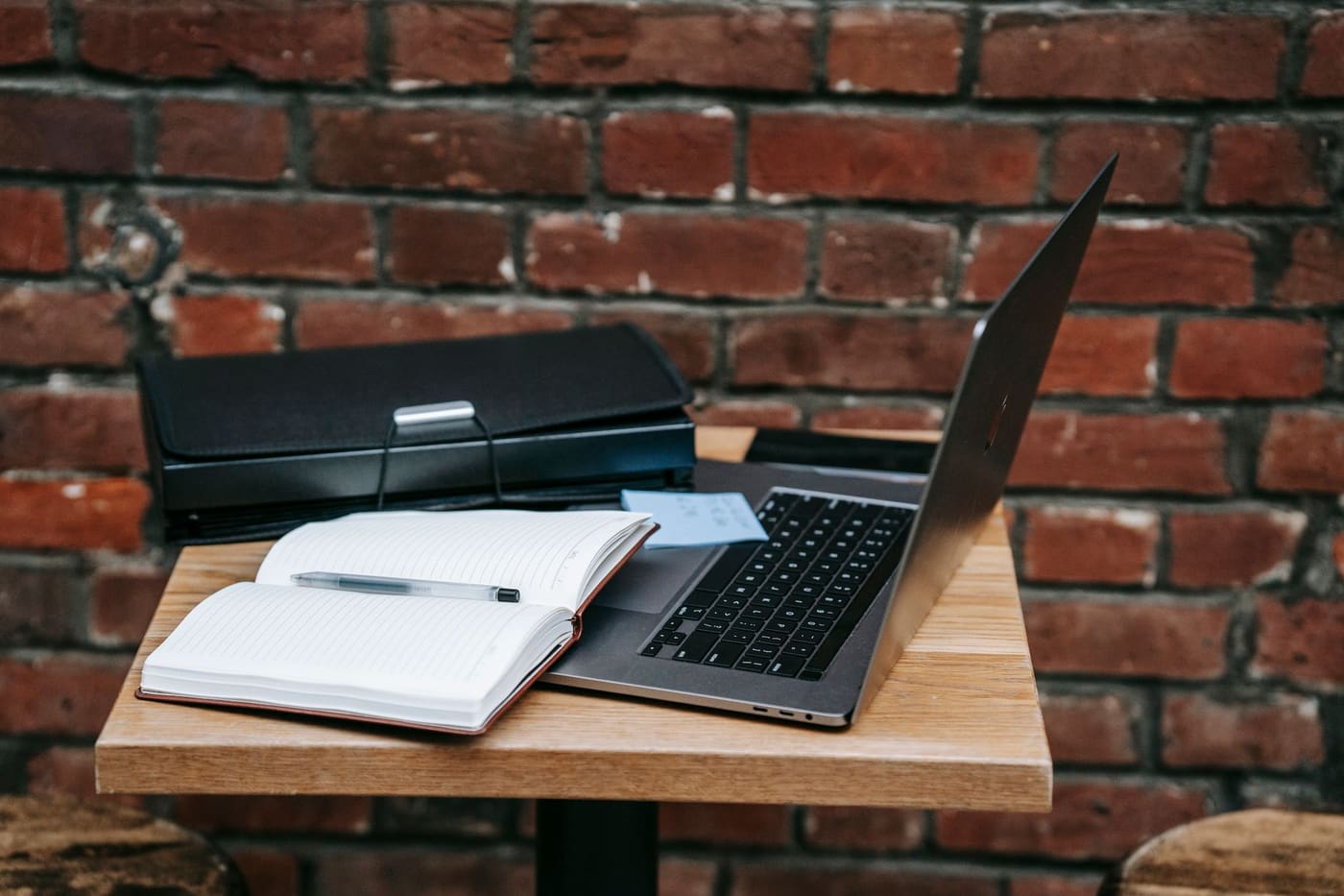 An open laptop and notebook on a small wooden table by a brick wall