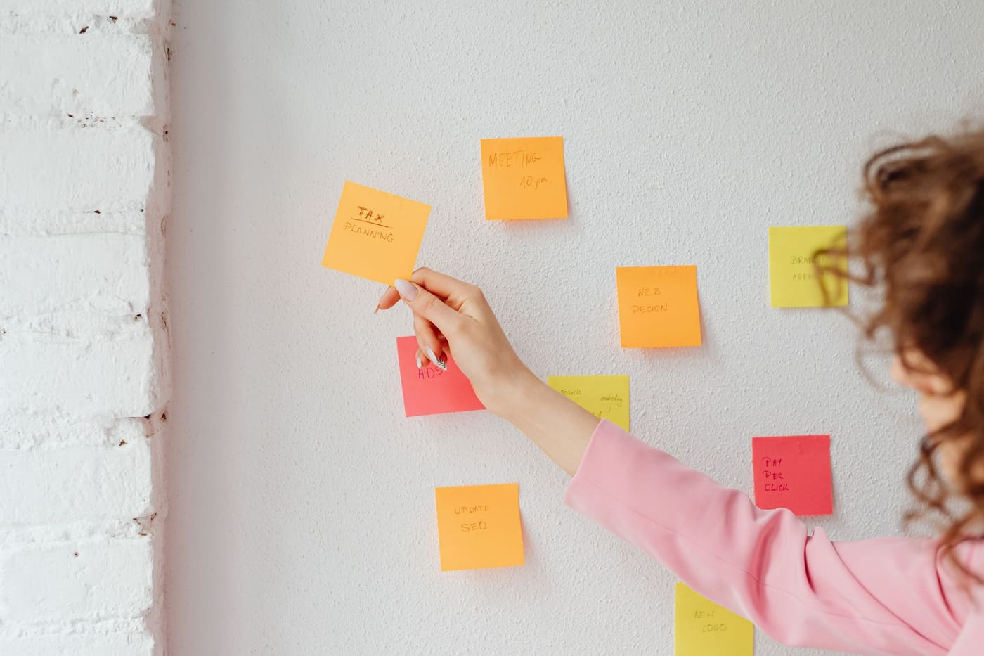 A woman arranging orange and pink sticky notes on a white wall to plan tasks
